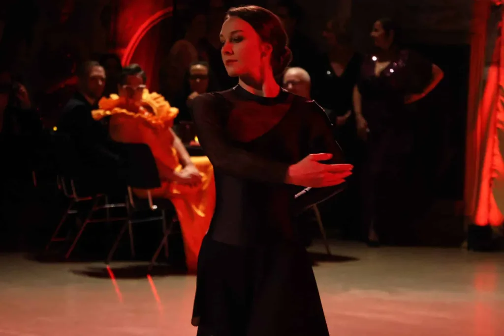 Eine Frau in schwarzer Kleidung tanzt in einem rot beleuchteten Saal, während im Hintergrund Gäste sitzen und stehen.