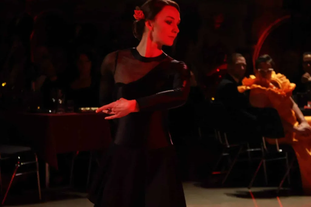 Tänzerin in schwarzem Kleid mit roter Blume im Haar bewegt sich bei gedämpfter roter Beleuchtung durch einen Saal.