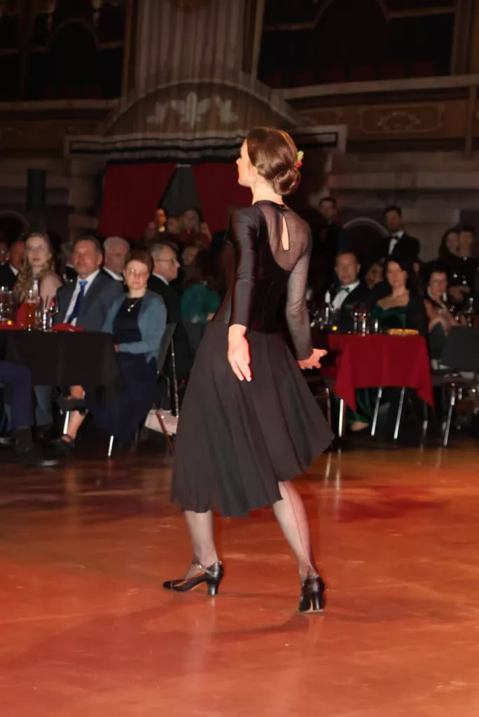 Tänzerin in schwarzem Kleid auf einer beleuchteten Tanzfläche vor sitzendem Publikum