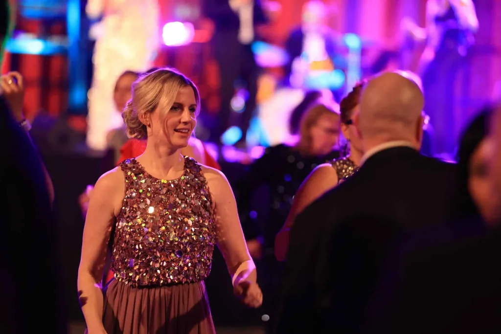 Eine Frau in einem glitzernden ärmellosen Kleid tanzt in einem farbenfroh beleuchteten Raum, während weitere Menschen im Hintergrund stehen.