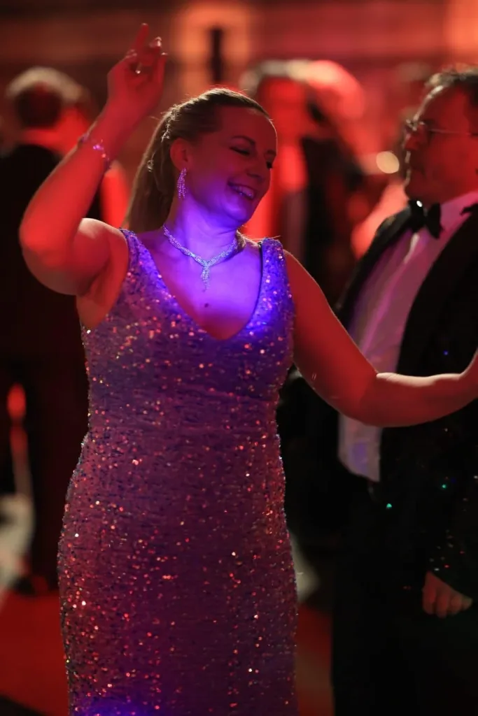 Tanzende Frau in einem glitzernden Kleid hebt einen Arm; neben ihr steht ein Mann im Smoking.