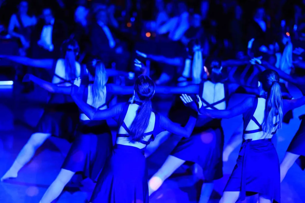 Mehrere Frauen tanzen in einer Gruppe unter blauem Licht; einige tragen dunkle Kleider mit hellen Tüchern.