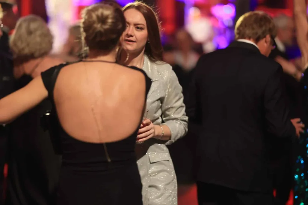 Mehrere elegant gekleidete Gäste tanzen in einem festlich beleuchteten Raum; im Vordergrund eine Frau mit hochgestecktem Haar und schwarzem Kleid, dahinter eine Frau im silberfarbenen Hosenanzug.