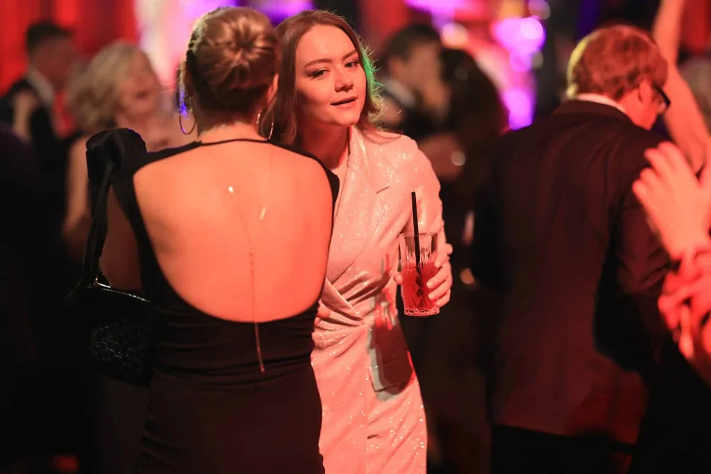 Eine Frau in glitzerndem Kleid hält ein rotes Getränk, während vor ihr eine weitere Frau tanzt.