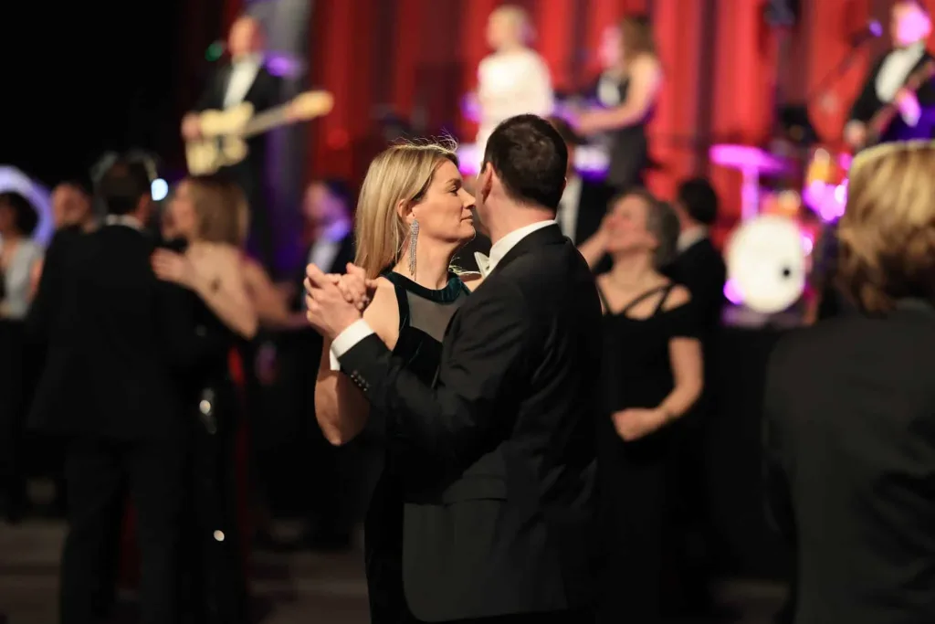 Ein elegant gekleidetes Paar tanzt im Vordergrund, während im Hintergrund weitere Menschen und eine Liveband zu sehen sind.