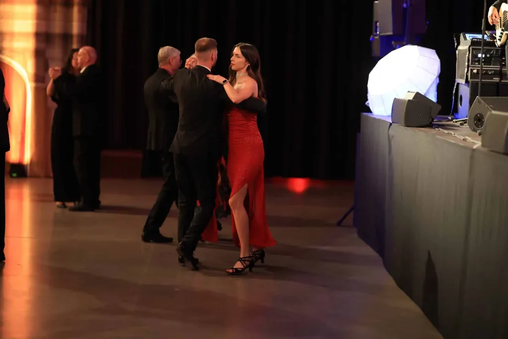 Ein Paar tanzt in eleganter Abendgarderobe auf einer beleuchteten Tanzfläche; rechts sind Bühne und Veranstaltungstechnik zu sehen.
