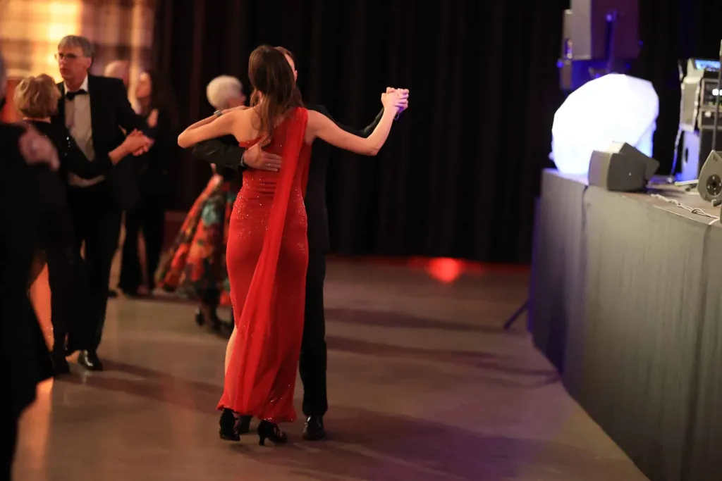 Ein Paar tanzt in einem roten Kleid auf einer festlich beleuchteten Tanzfläche; weitere Tanzende und eine technische Ausstattung sind im Hintergrund sichtbar.