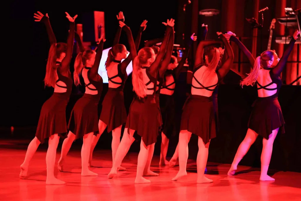 Eine Gruppe junger Tänzerinnen hebt auf einer rot beleuchteten Bühne die Arme zu einer gemeinsamen Choreografie.