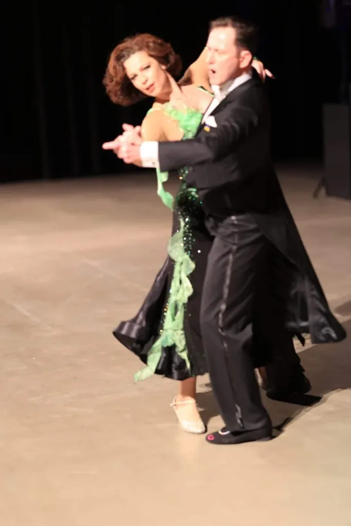 Ein Paar tanzt in eleganter Abendgarderobe über eine beleuchtete Tanzfläche. Die Frau trägt ein grünes, glitzerndes Kleid, der Mann einen schwarzen Frack.