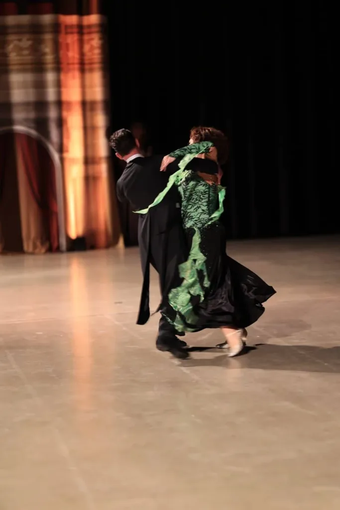 Ein Tanzpaar bewegt sich auf einer Bühne; die Person im Vordergrund trägt ein schwarzes Kleid mit grünen Verzierungen.