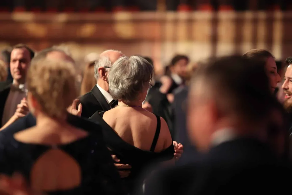 Mehrere elegant gekleidete Tanzpaare bewegen sich auf einer dicht besetzten Tanzfläche; im Mittelpunkt tanzt eine Frau mit kurzem grauem Haar und schwarzem Kleid mit einem Mann im dunklen Anzug.