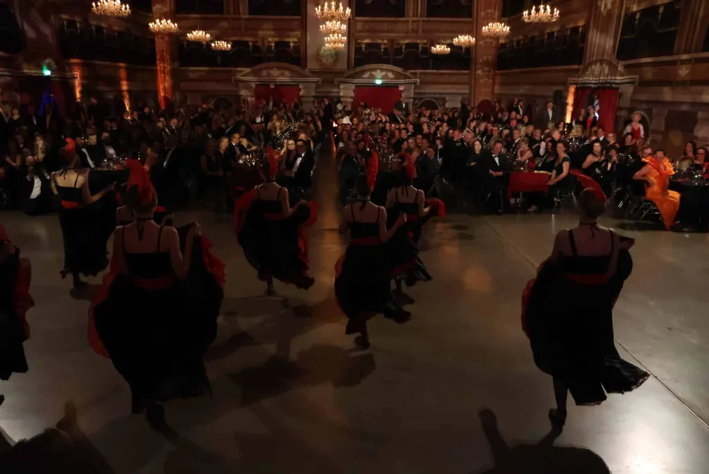 Tanzende in schwarzen Kleidern mit roten Akzenten führen in einem festlich beleuchteten Saal eine Choreografie vor, während das Publikum zuschaut.