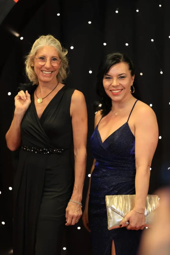 Zwei lächelnde Frauen stehen vor einem schwarzen, mit Lichtpunkten besetzten Hintergrund. Eine trägt ein schwarzes Kleid, die andere ein dunkelblaues Kleid und hält eine goldfarbene Clutch.