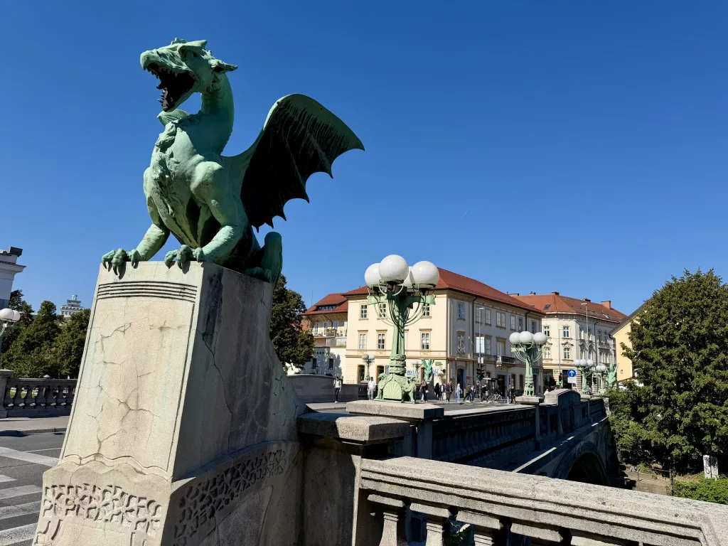 Grüne Drachenstatue auf einem hohen steinernen Sockel vor historischen Gebäuden und einem klaren blauen Himmel