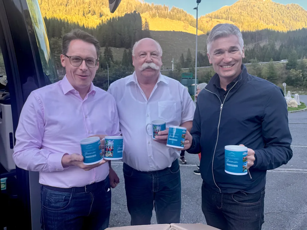 Drei Männer stehen im Freien vor einer Berglandschaft und halten bedruckte Becher in die Kamera.