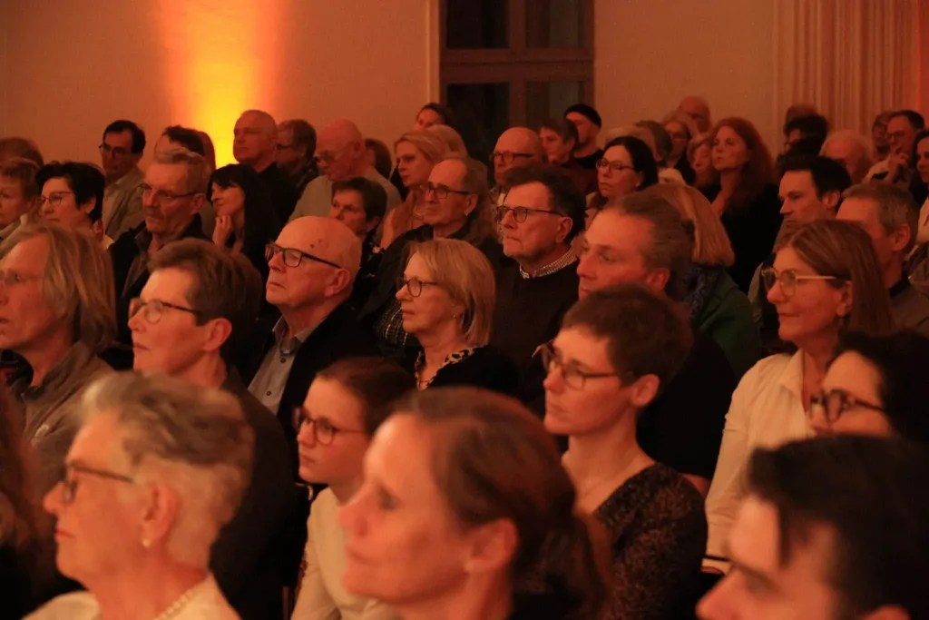 Dicht stehendes und sitzendes Publikum bei einer Veranstaltung in warm beleuchtetem Innenraum