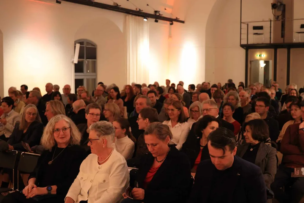 Dicht sitzendes Publikum verfolgt eine Veranstaltung in einem festlich beleuchteten Saal.