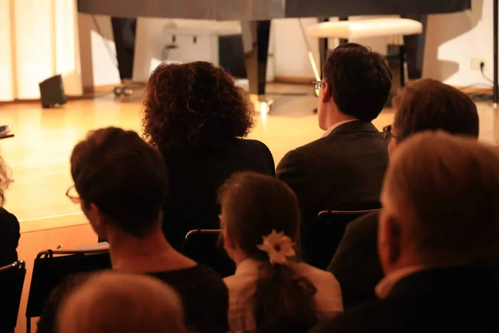 Publikum sitzt in einem Saal und blickt auf eine beleuchtete Bühne.