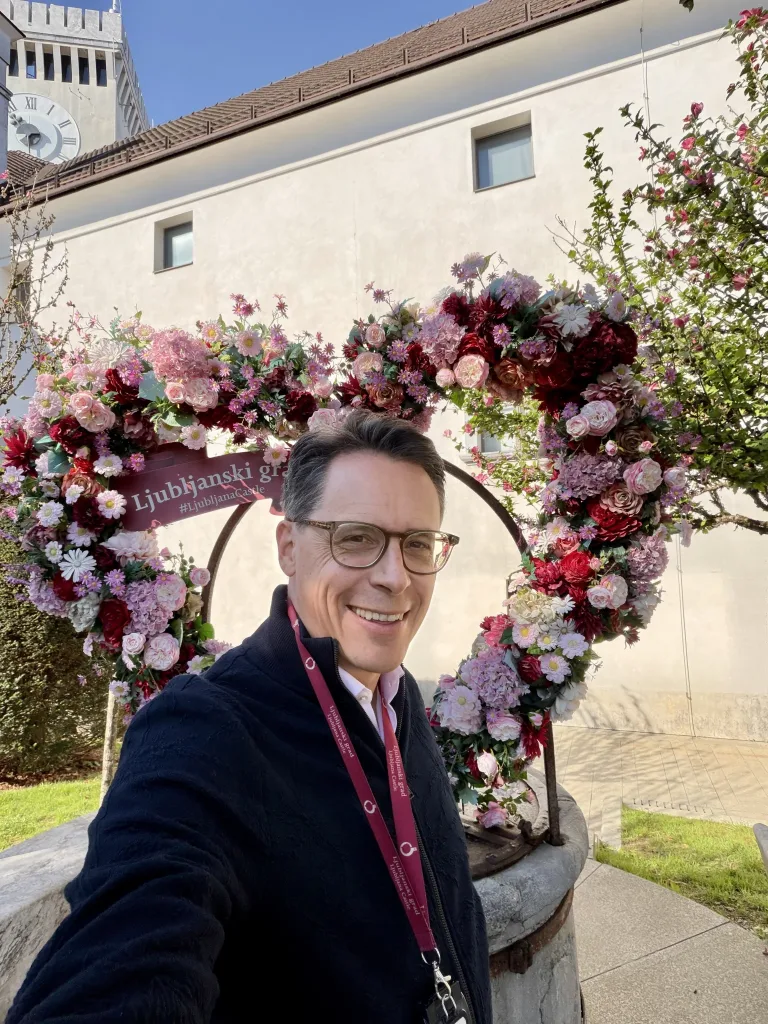 Mann mit Brille und dunkler Kleidung steht vor einem großen, farbenfrohen Blumenbogen