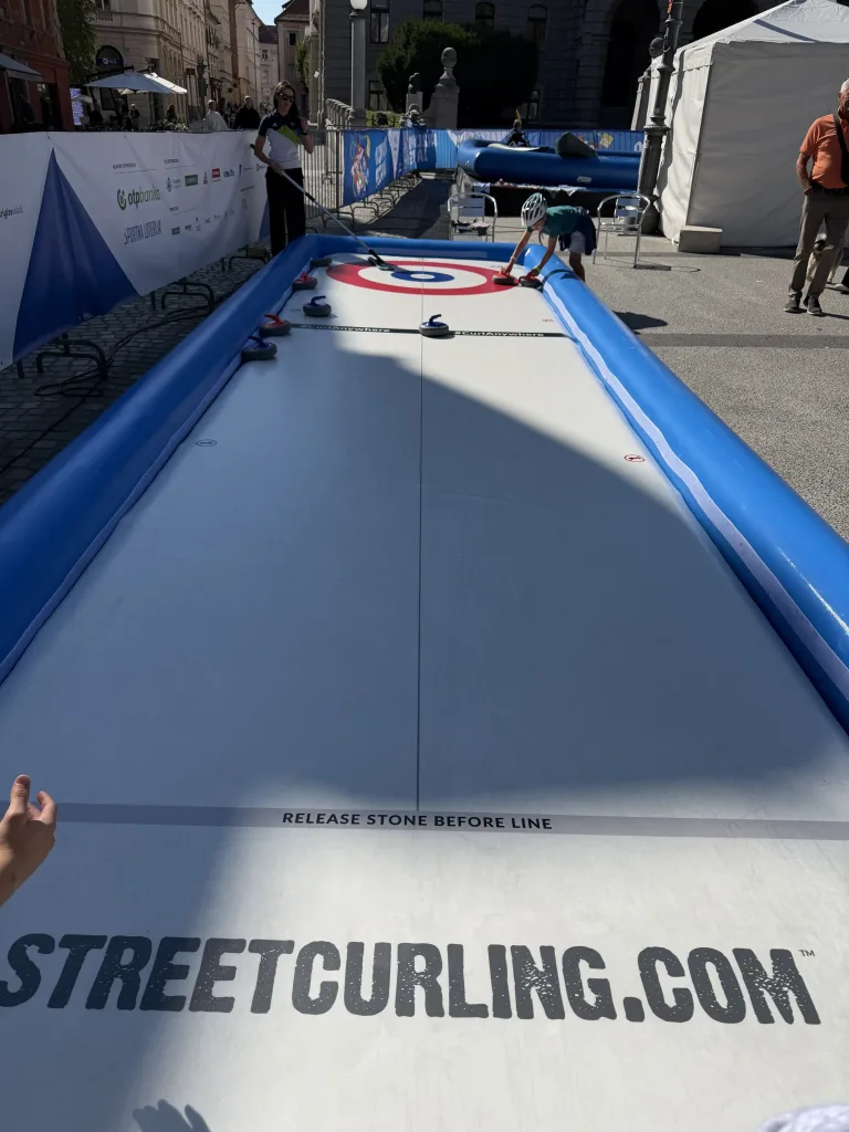 Mobile Curlingbahn im Freien; eine Person gibt einen Stein frei, eine weitere wischt die Bahn.