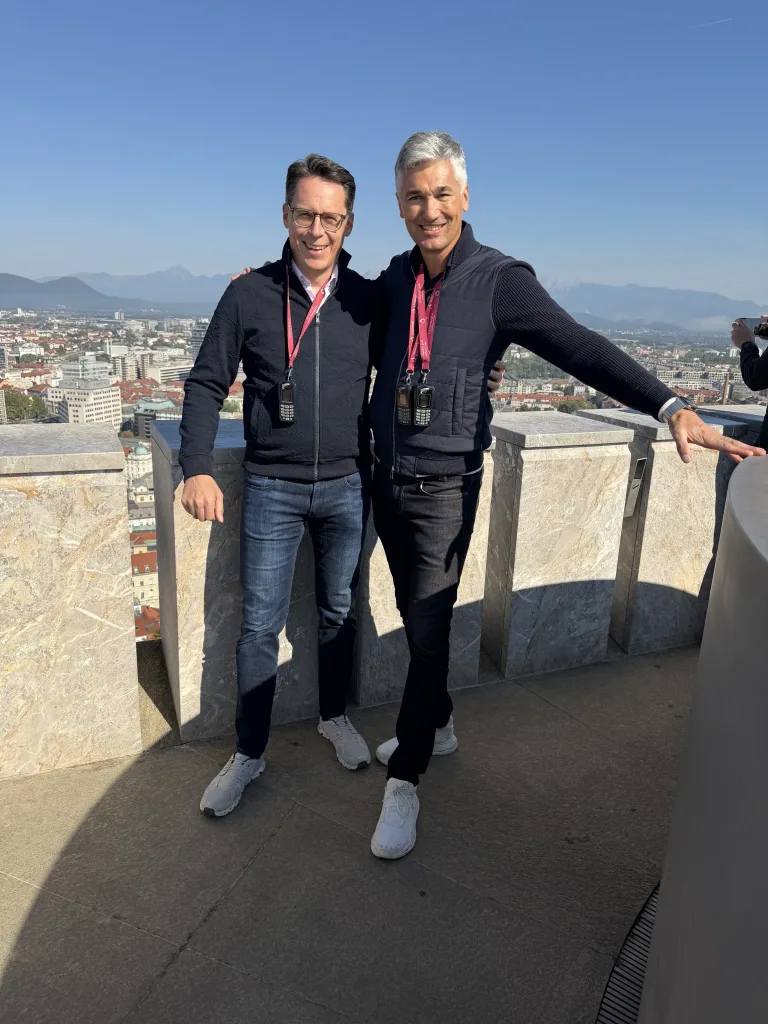 Zwei Männer stehen auf einer Aussichtsterrasse vor einem weiten Stadt- und Bergpanorama.